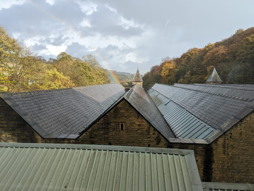 A rainbow drops between the spires above the weaving shed roofs